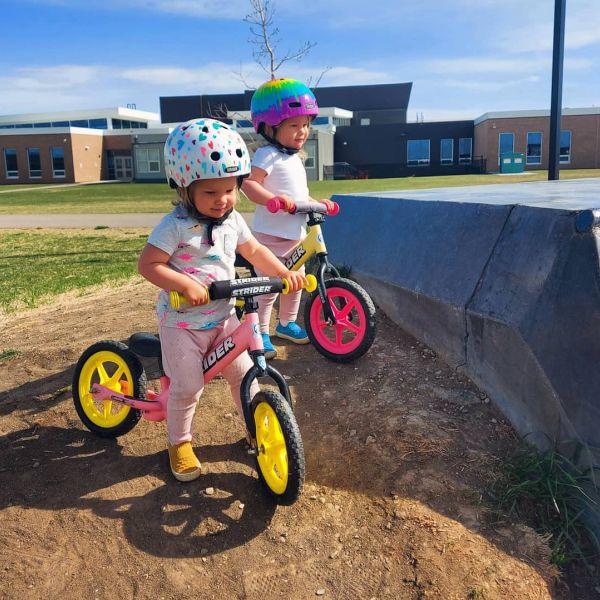 Two toddlers wearing matching light pink pants, t-shirts, and helmets stride on their colorful, custom Strider Bikes on a sunny day. One girl is riding a pink bike with yellow Ultralight Wheels and yellow grips. The other girl is riding a yellow bike with hot pink Ultralight Wheels and pink grips.