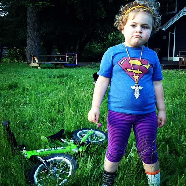 Girl in Superwoman shirt and mismatched socks poses in front of green Strider bike that's laying in the grass