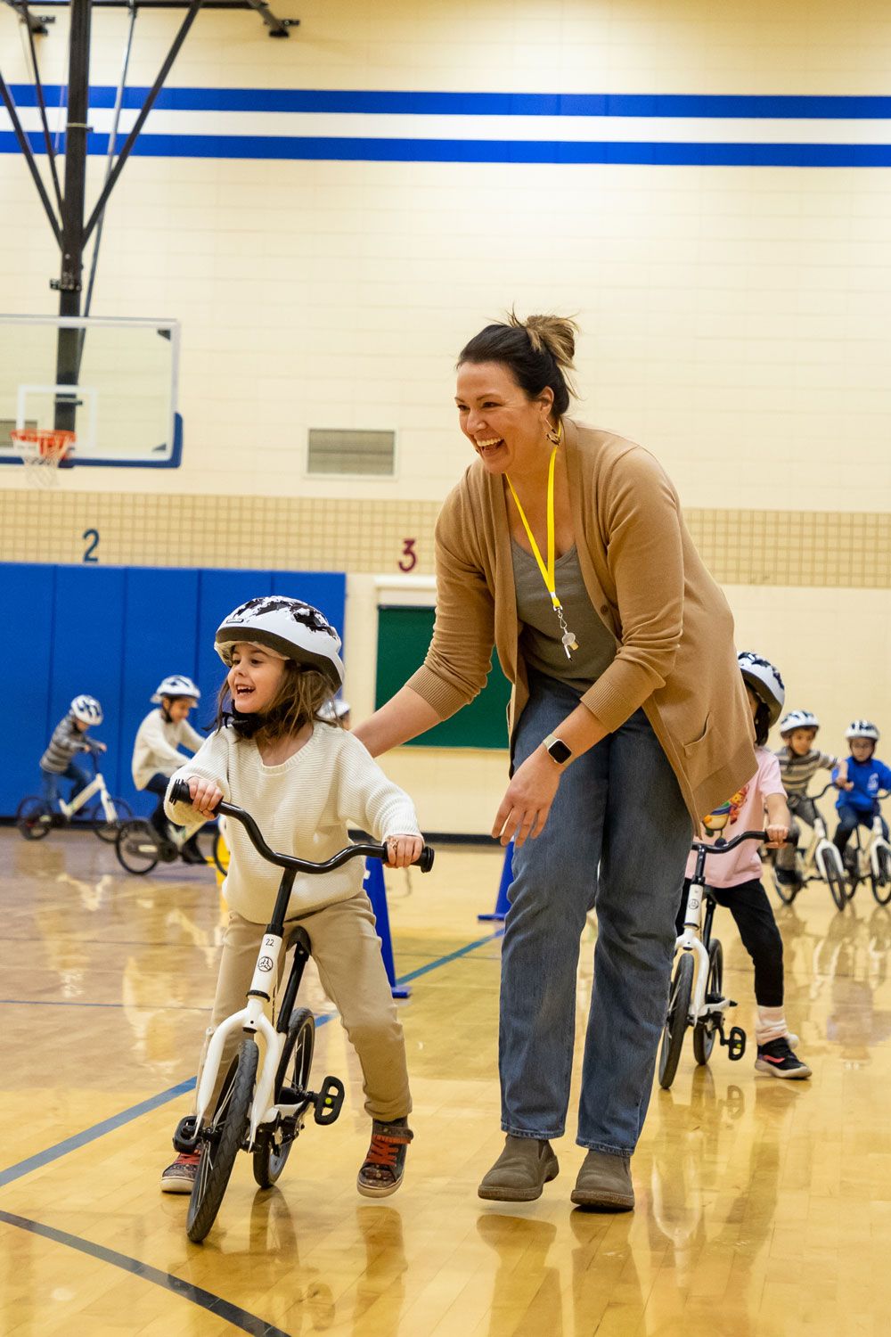 A teacher smiles while helping a girl learn to pedal in the All Kids Bike Kindergarten PE Program