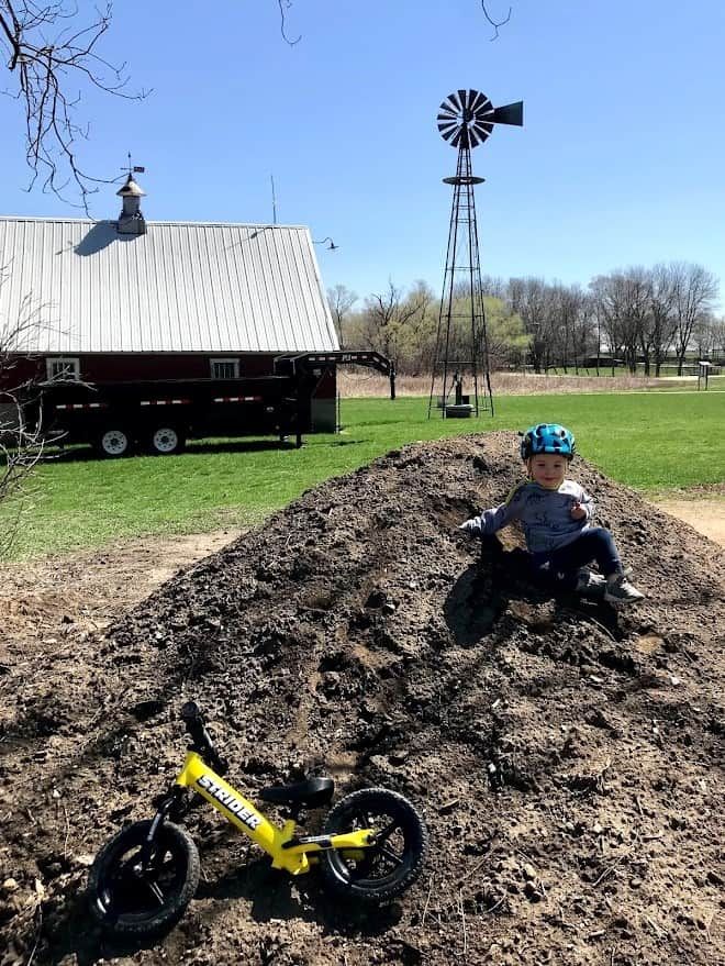Owen Tudor sits on a dirt pile on a farm with his yellow Strider bike in the foreground.