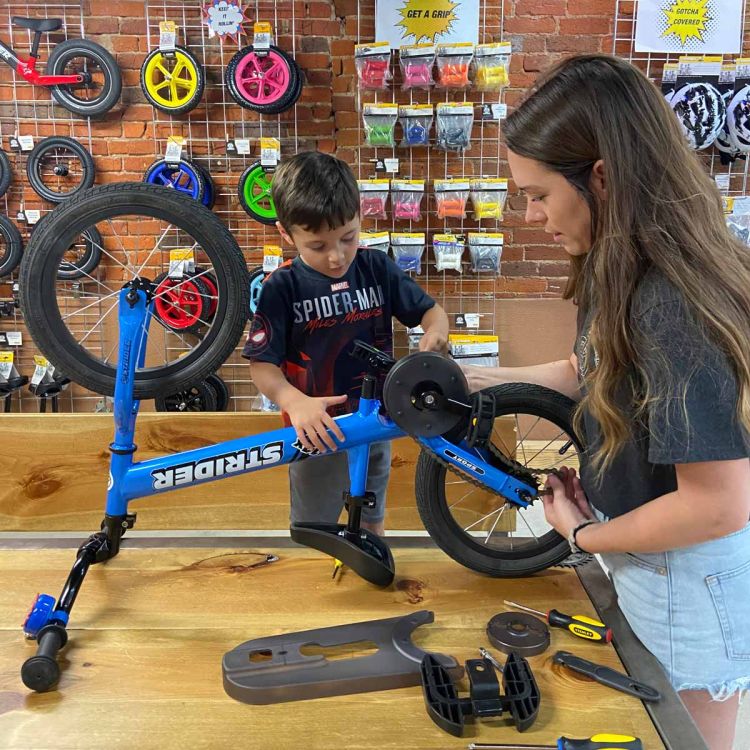 A boy works on the pedals of his Strider Bike while an adult supervises