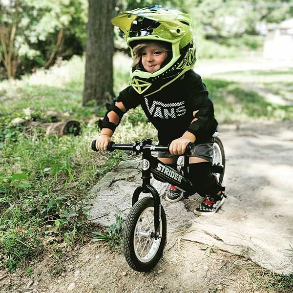 Child in a full-face helmet, Vans shirt and checkered shoes riding a tricked-out Strider balance bike
