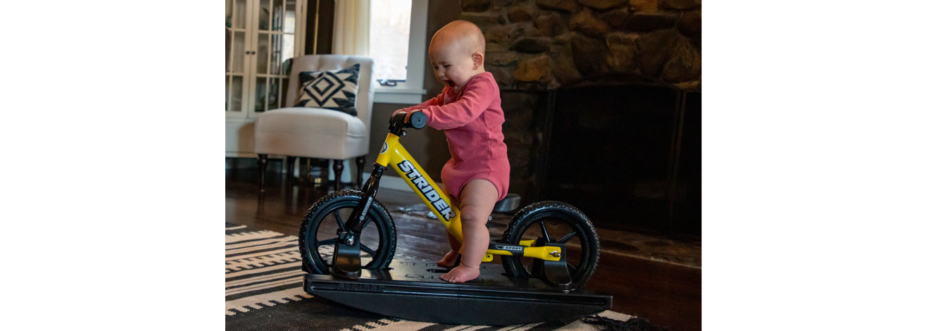 A baby riding on a yellow Strider 2-in-1 Rocking Bike in the living room