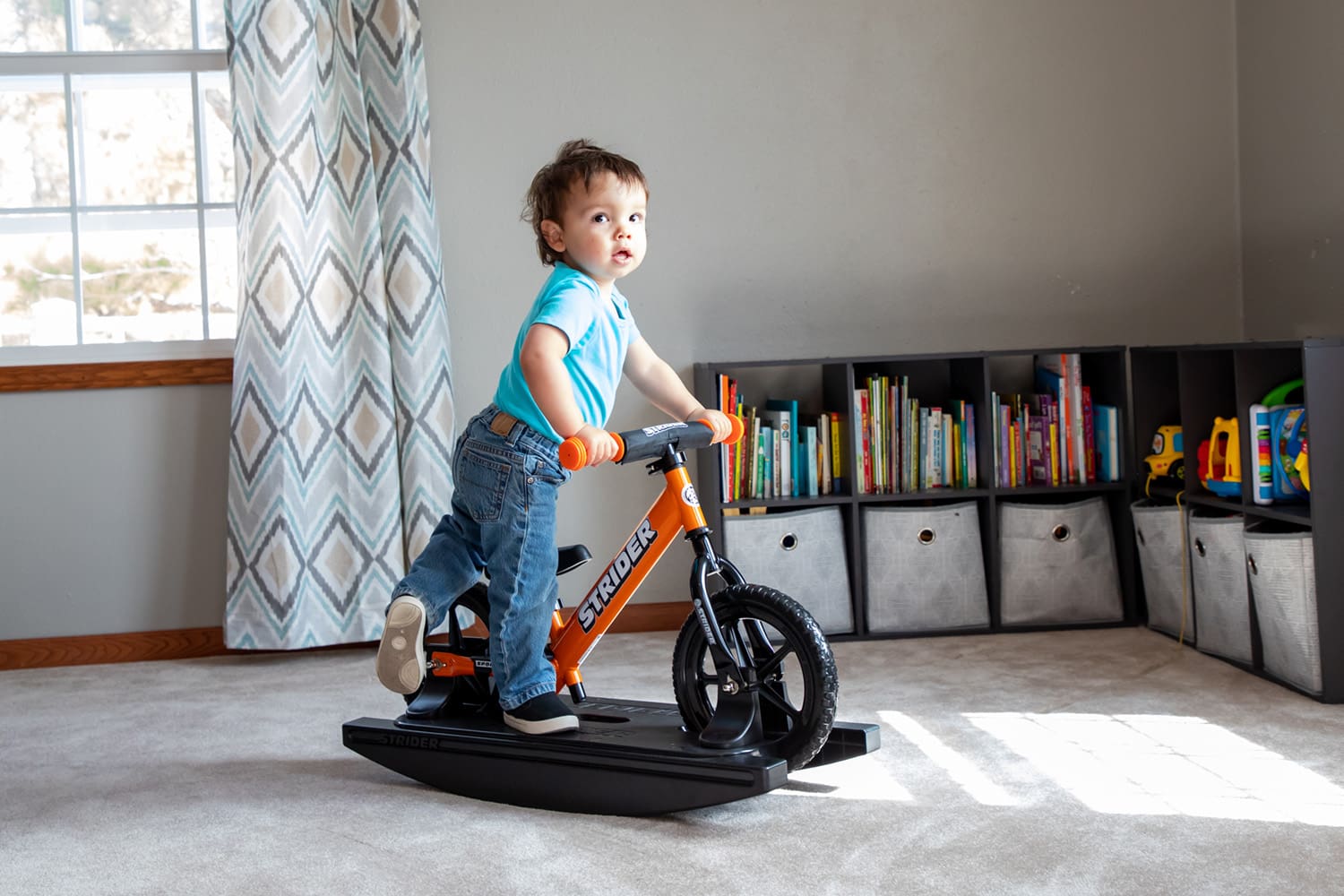A toddler steps onto an orange Strider Rocking Bike