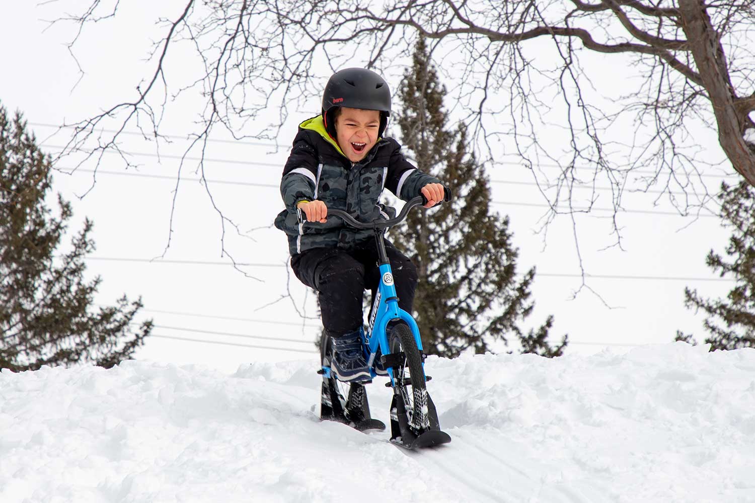 An excited boy rides downhill on a Strider bike with ski attachments
