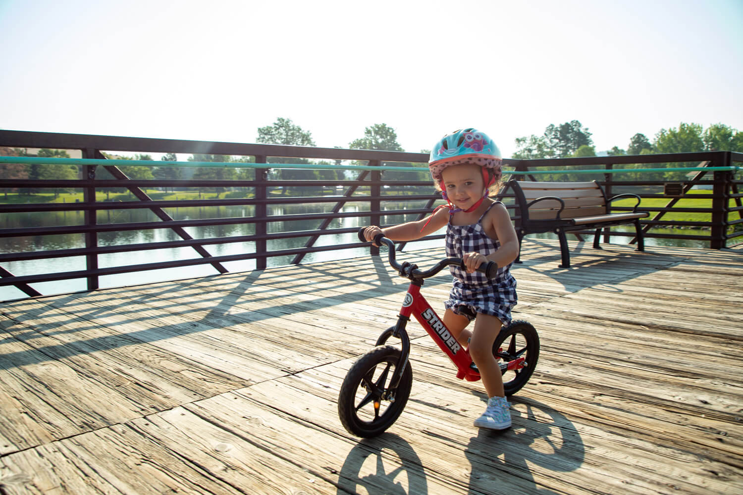 A girl on a red balance bike with Strider High Rise Wide Handlebars rides a wooden path next to a lake