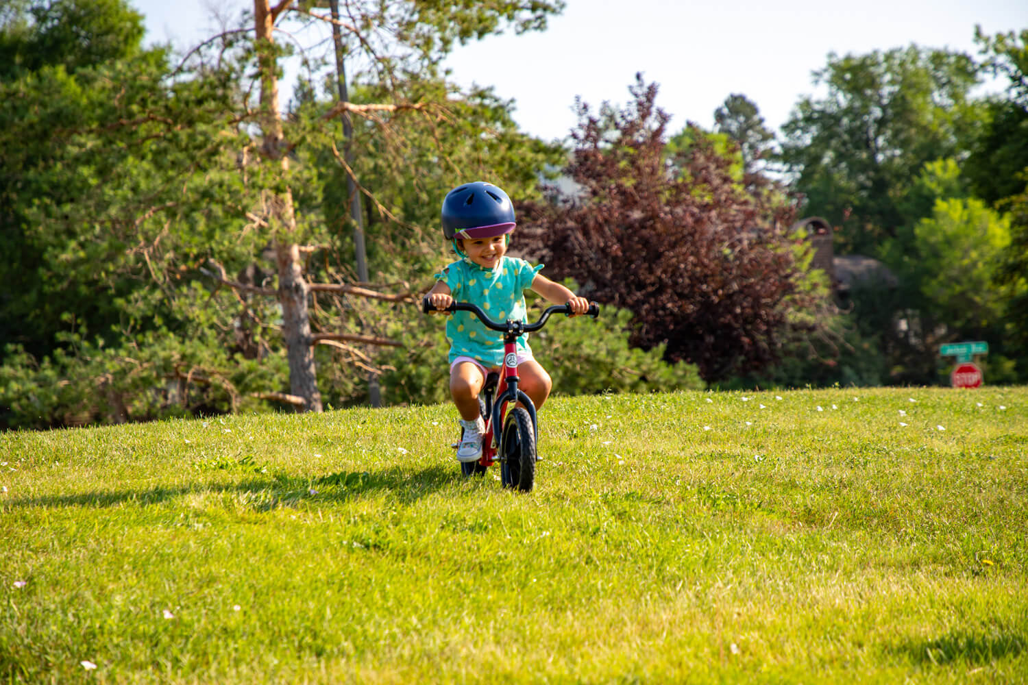 A girl on a red balance bike with Strider High Rise Wide Handlebars rides down a grassy hill