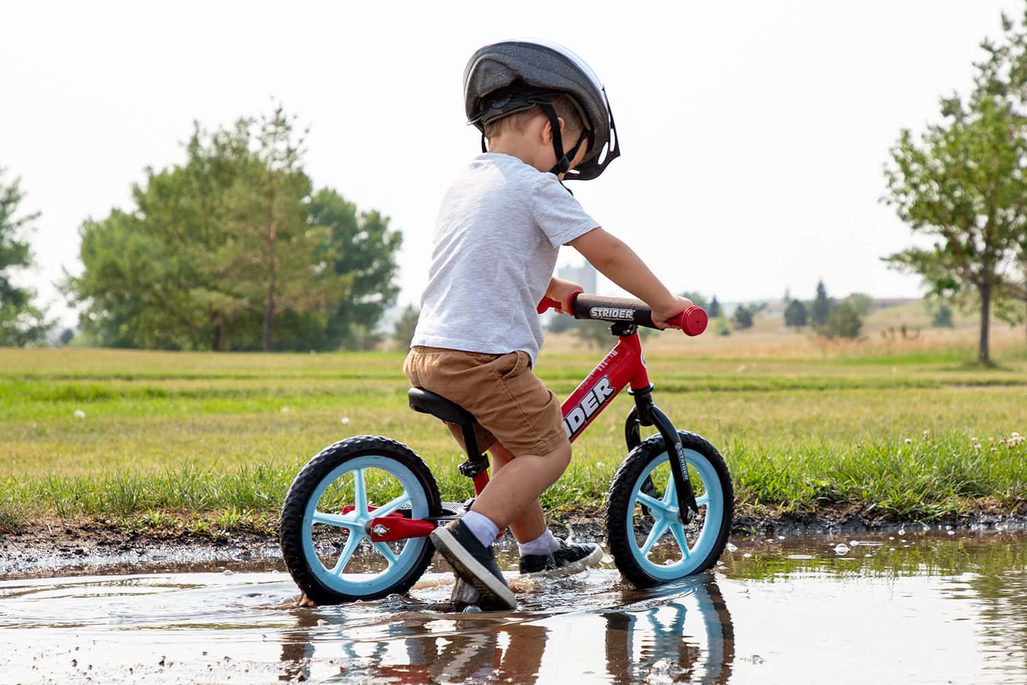 A young boy rides through a puddle in a park on a red Strider 12 Sport bike with light blue Ultralight Wheels