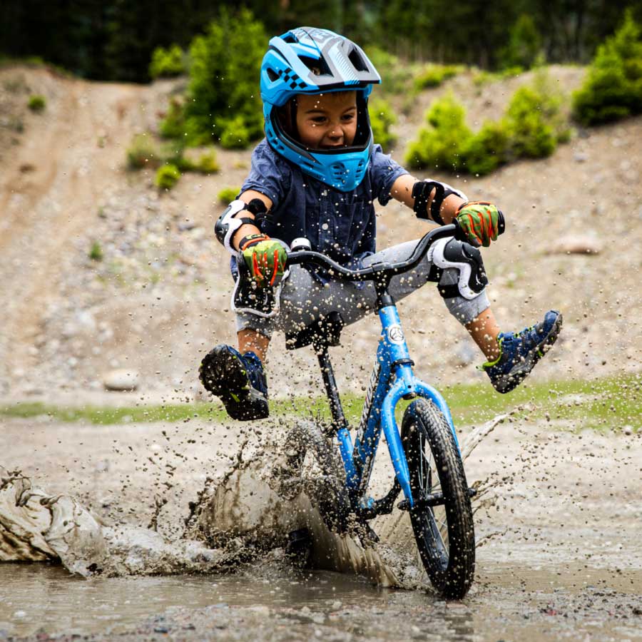 A boy rides a blue Strider 14x through a mud puddle