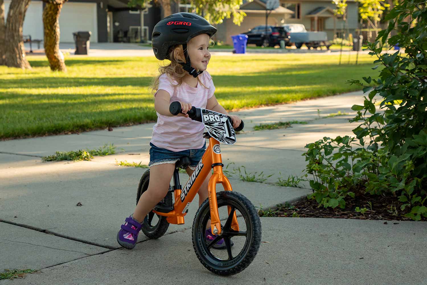 A girl riding an Orange Rush Strider 12 Pro balance bike through the neighborhood