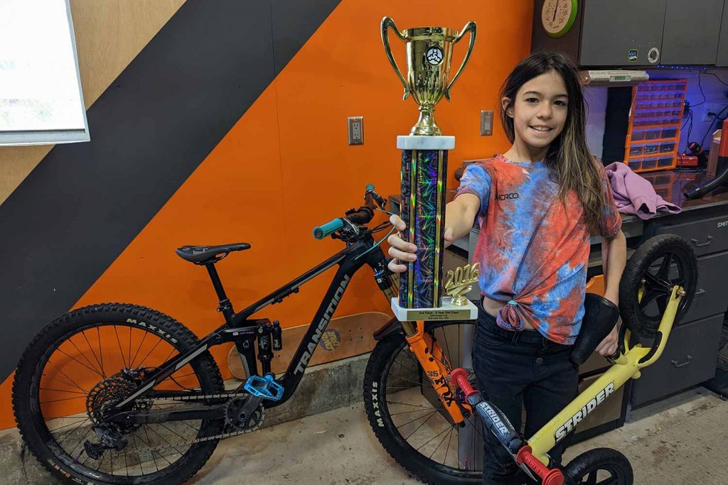 Sophie Gregory holding her first trophy, from a Strider Cup race, and posing with her old bike (and her new one in the background)